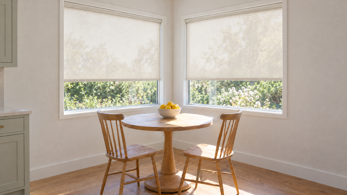 Light Filtering Roller Shades in Ivory — a breakfast nook in a family kitchen where two windows meet at an inside corner, late morning with bright directional sun