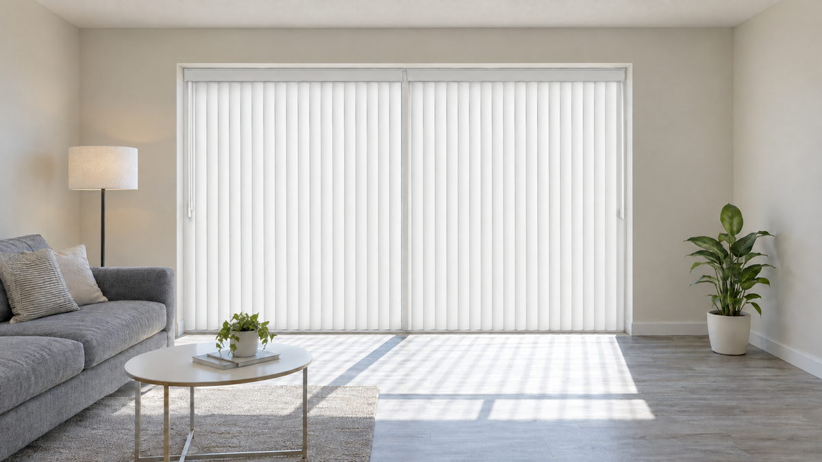 Vertical Blinds in White — a modest rental apartment living room, lightly furnished: a compact gray fabric sofa, a small round white coffee table, a floor lamp, a single potted plant, off-white builder-grade painted walls, light gray wood-look vinyl