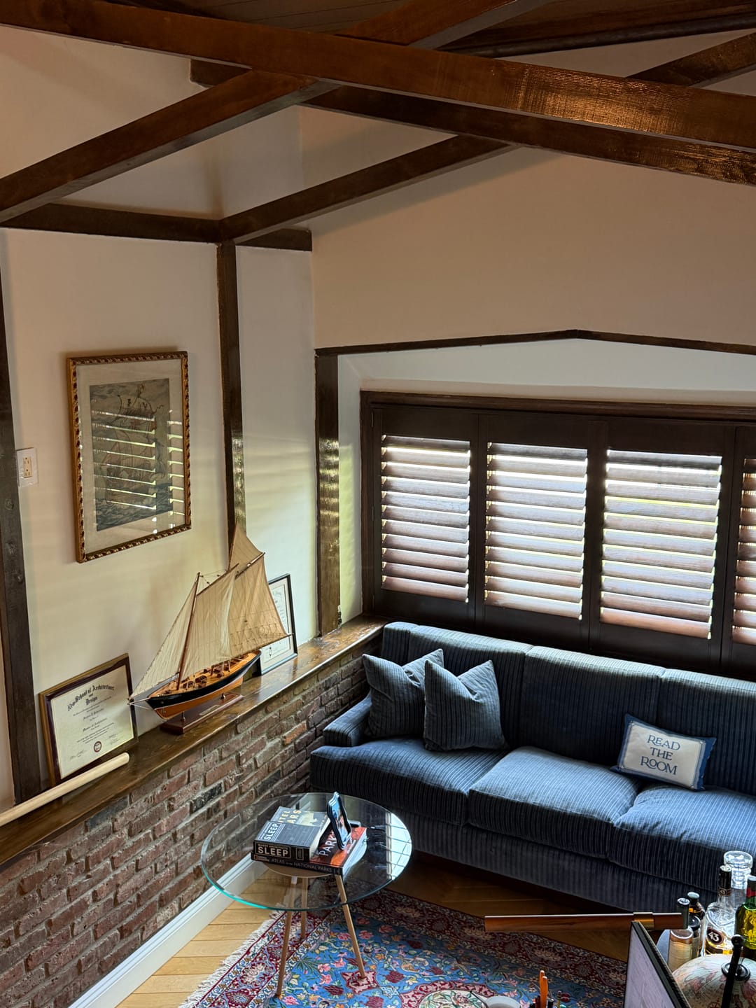 A cozy living room corner featuring a navy blue corduroy sofa with matching pillows, set against a brick wall and a window with dark wood shutters. A small wooden model ship sits on the windowsill, and a glass-top side table is placed in front of the couch.