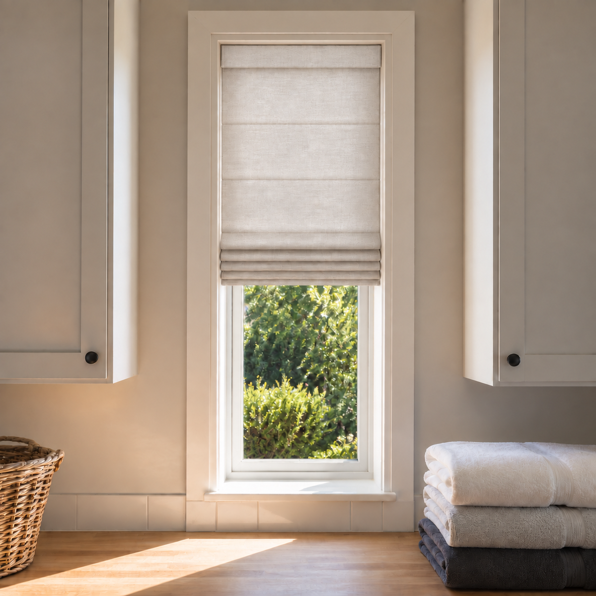 A light beige fabric Roman blind partially drawn on a tall window, set in a bright, modern laundry room with white cabinets, a wooden floor, and folded towels.